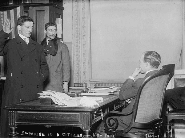 Photo: a man taking the required citizenship oath of allegiance in front of a judge in New York City (1910). Credit: Library of Congress, Prints and Photographs Division.