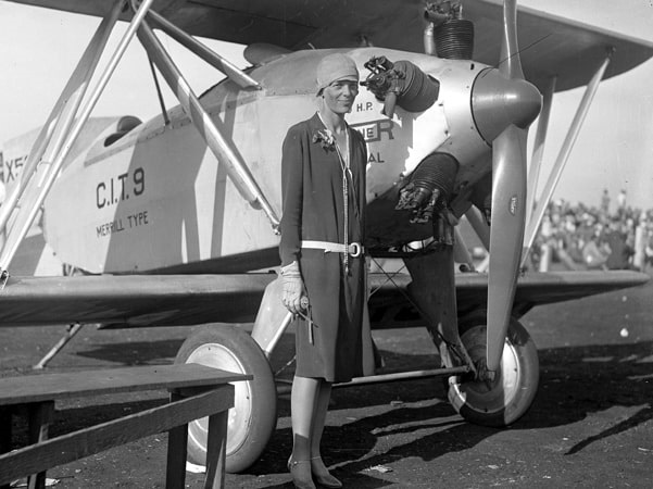 Photo: Amelia Earhart, wearing a dress, standing beside a Merrill CIT-9 Safety Plane, c. 1928. Credit: UCLA Library; Wikimedia Commons.