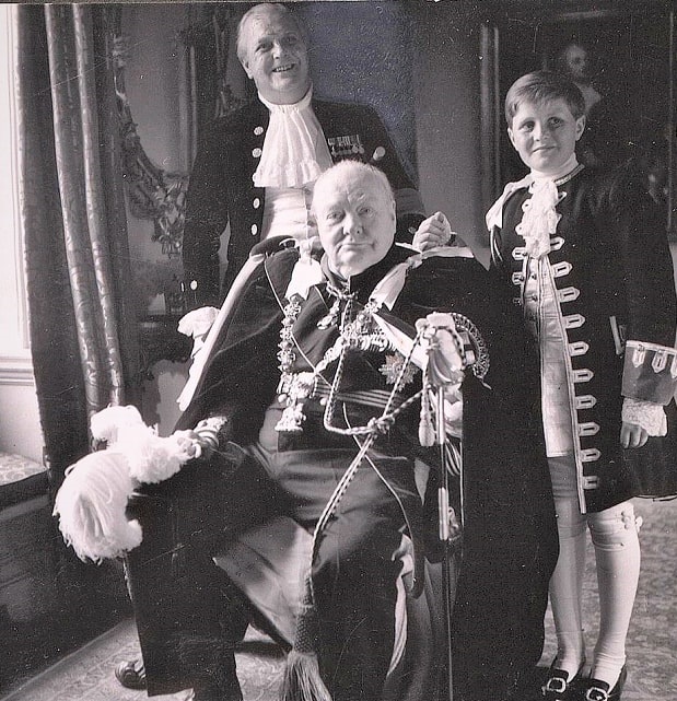 Photo: three generations of Churchills: Sir Winston Leonard Spencer Churchill (seated) with son Randolph Frederick Edward Spencer Churchill (1911-1968) and grandson Winston Spencer Churchill (1940-2010) [the son of Randolph Frederick Edward Spencer Churchill and Pamela Digby] wearing coronation robes of the Order of the Garter for the coronation of Queen Elizabeth II. Credit: Wikimedia Commons.