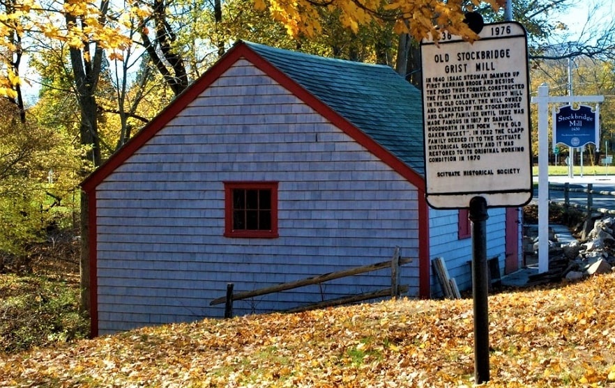 Photo: Old Stockbridge Grist Water Mill with historic marker, which reads: “In 1640 Isaac Stedman dammed up First Herring Brook and beside the pond thus formed, constructed the first water driven grist mill in the old colony. The mill, owned and operated by the Stockbridge and Clapp families until 1922, was made famous in 1817 by Samuel Woodworth in his poem ‘The Old Oaken Bucket.’ In 1922 the Clapp family deeded it to the Scituate Historical Society and it was restored to its original working condition in 1970. Marker erected in 1976 by the Scituate Historical Society in Scituate, Massachusetts. Courtesy of the Historical Marker Database.