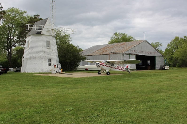 Photo: windmill, plane, and hangar at Cape Cod Airfield in Marstons Mills, Massachusetts, 26 May 2013. The airport was founded by Zenas Crocker, Katie Crocker’s great uncle. It opened with a flying circus on 4 July 1929. Credit: Kevin Rutherford; Wikimedia Commons.