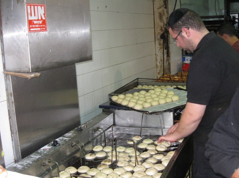 Photo: a baker deep-fries sufganiyot at the Mahane Yehuda Market (shuk) during Hanukkah. Credit: Yoninah; Wikimedia Commons.