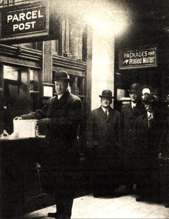 Photo: Postmaster General Frank Hitchcock mailing a package to Postmaster Morgan. A group of postal officials, including Chief Clerk George Thompson (middle) watch. Credit: Smithsonian National Postal Museum.